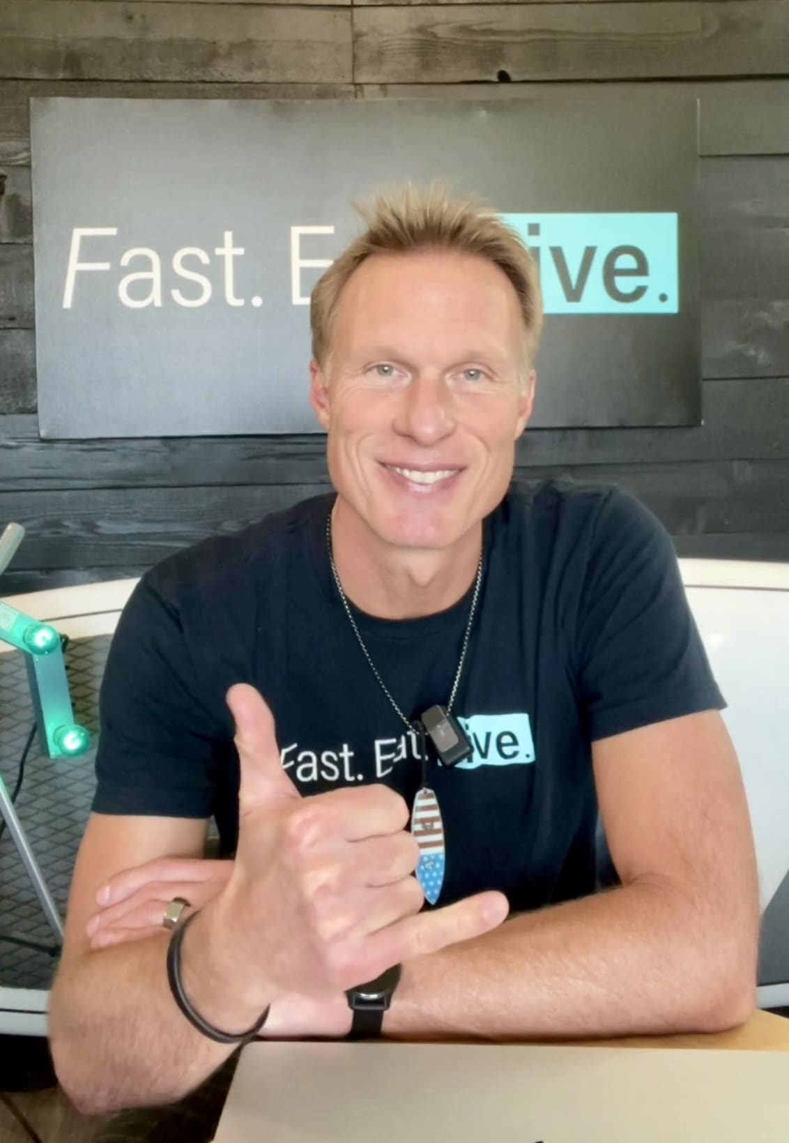 Man with short blonde hair wearing a "Fast. Effective." shirt and matching sign, smiling and making a shaka hand gesture