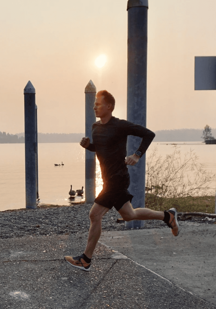 Man running outdoors near water during sunset with a calm sky and several poles in the background.